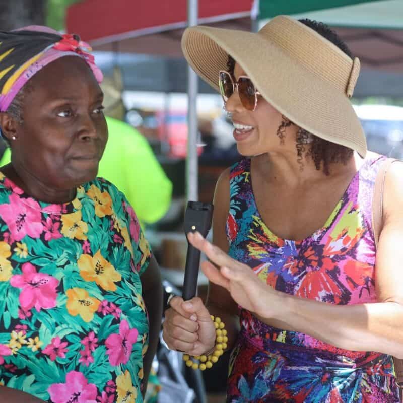 Two women outdoors, one wearing a floral print dress and sun hat, holding a microphone and speaking animatedly to the other, who is wearing a colorful floral top and headscarf at the King of the Wing contest in Magens Bay, St. Thomas, USVI.
