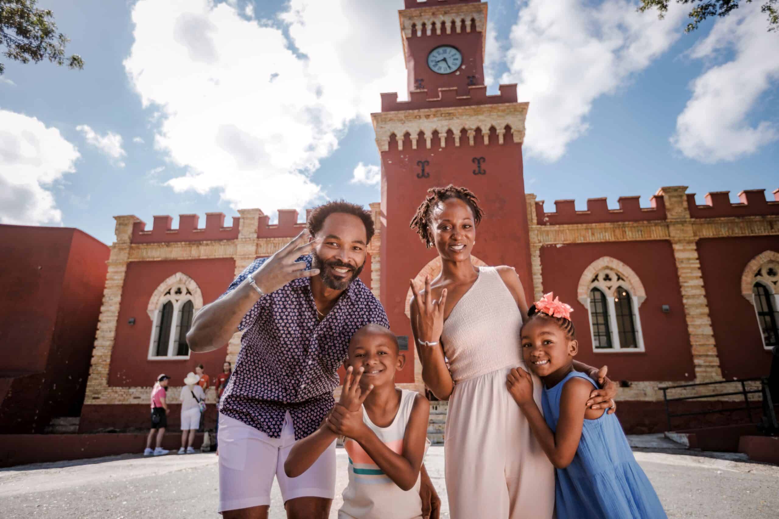 A family smiles and makes VI with their hands in front of Fort Christian on St. Thomas, USVI