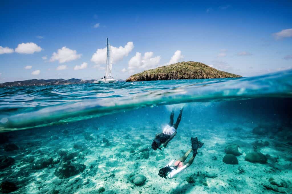 Above the water, a sailboat is floating by an island, below the surface, two people are snorkeling through coral off of Buck Island, St. Croix