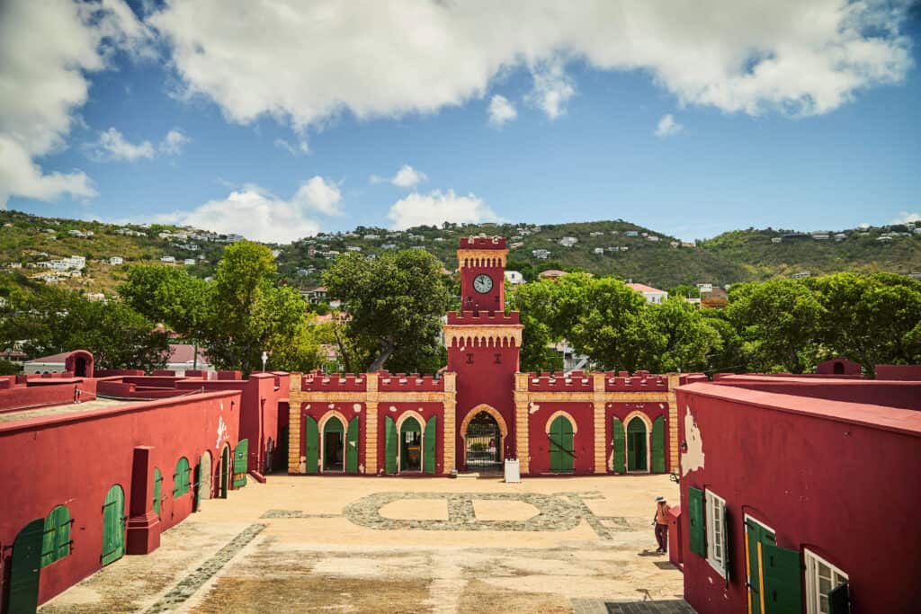 Fort Christian in Charlotte Amalie, St. Thomas, USVI features massive stone walls, tall widows with green shutters and a clocktower. Behind, there are verdant hills.