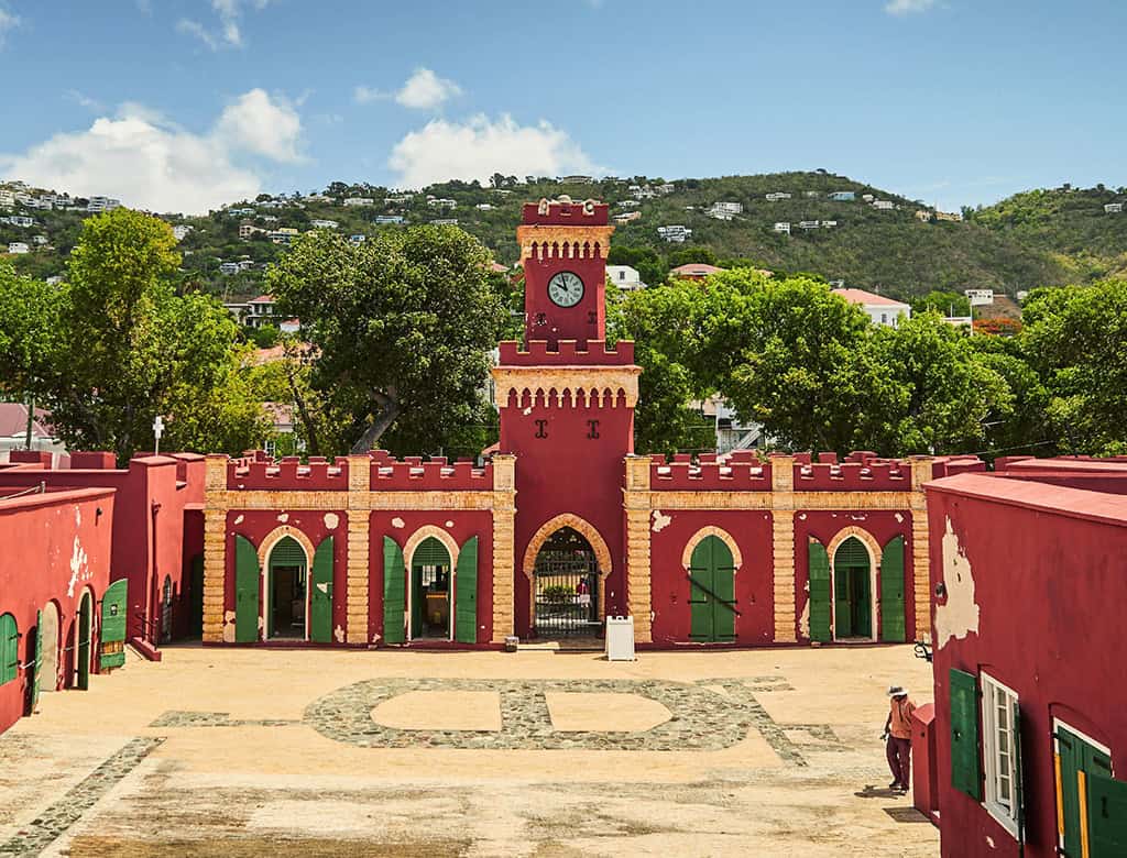 A tall, red fort has tall doors and a clean courtyard. Green hills are visible beyond the fort.