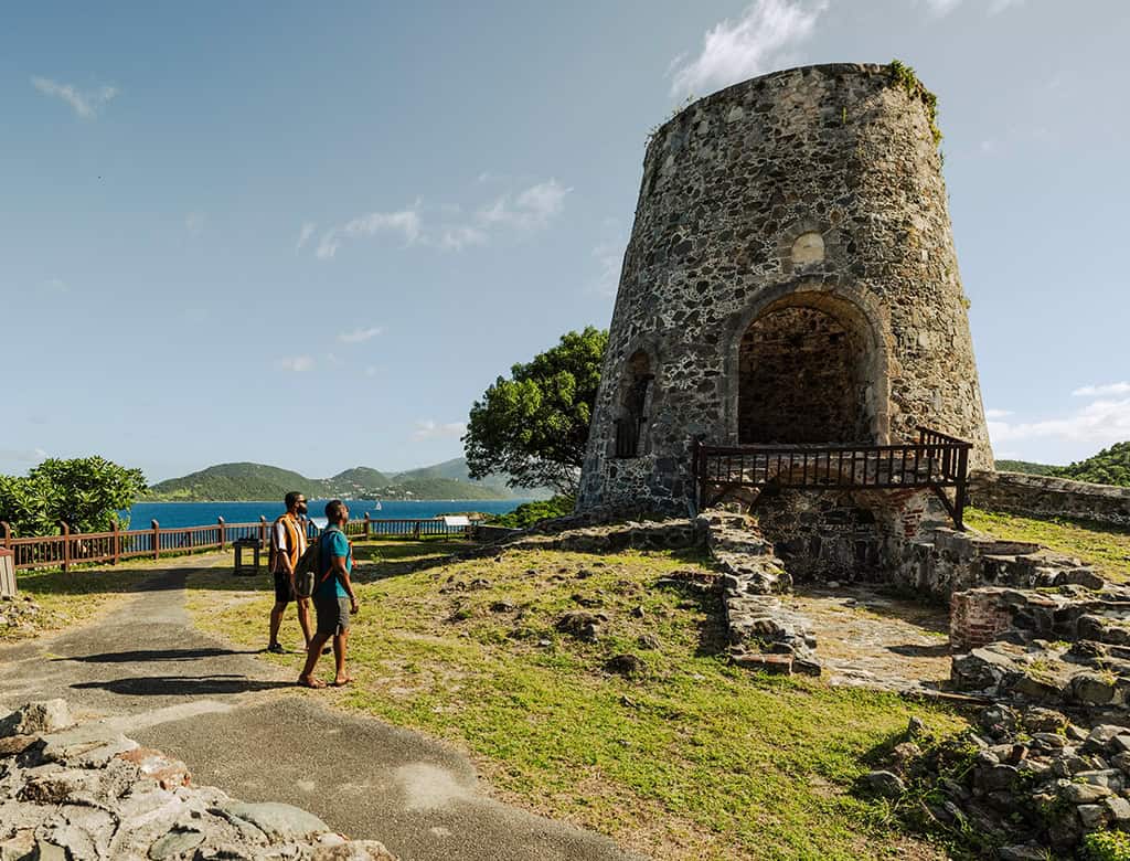 Two people walk on a path outside of a plantation ruin in USVI, the ocean and rolling hills are visible in the background.