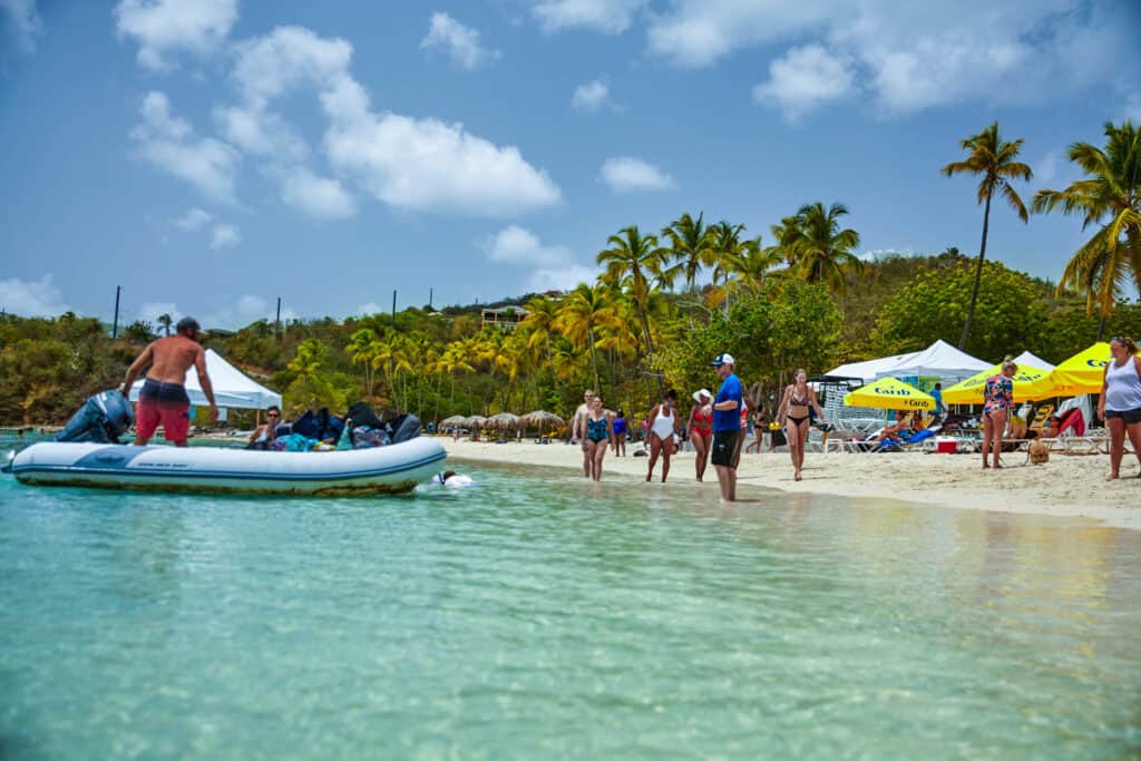 A group pulls up on a dingy as they arrive to a beach on Water Island. People walk along the waters edge and lounge on the sand