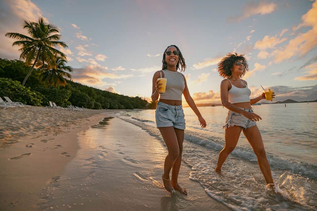 Two people smiling as they walk along the shore of Honeymoon Beach, St. John, USVI with drinks in hand