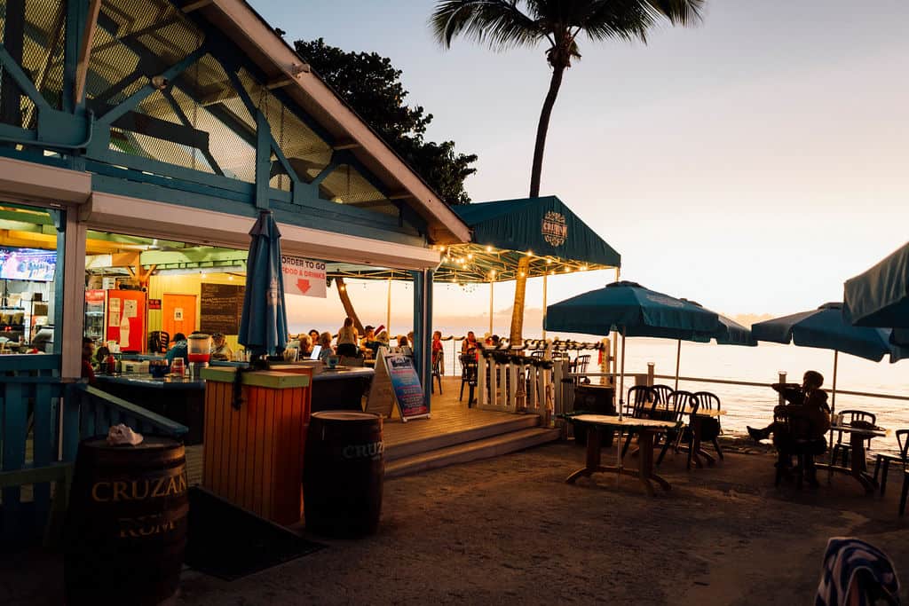 A beach bar in St. John glows at sunset. People are milling around on the deck sipping drinks and chatting.