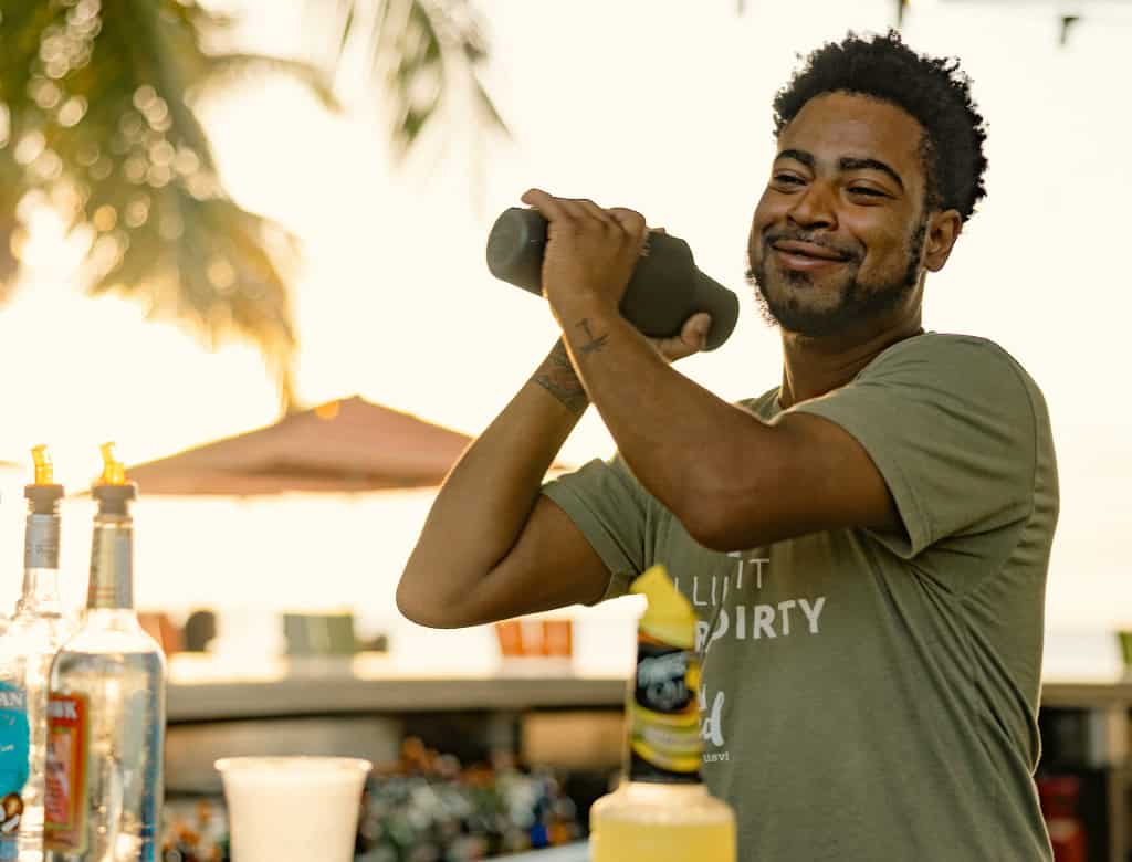 A bartender smiles as he shakes a cocktail in a shaker. Mixers are in the foreground and palm tress are in the background