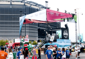 People gather at an outdoor event near a stadium, with a stage and banners displaying sponsors.