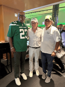 Three men are posing together indoors, one wearing a green New York jersey, another in a white outfit and cap, and the third in a white shirt and cap.