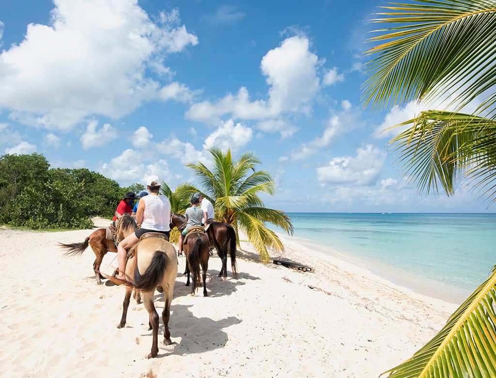 A group of people horseback ride along a white-sand beach with some white puffy clouds in the sky.