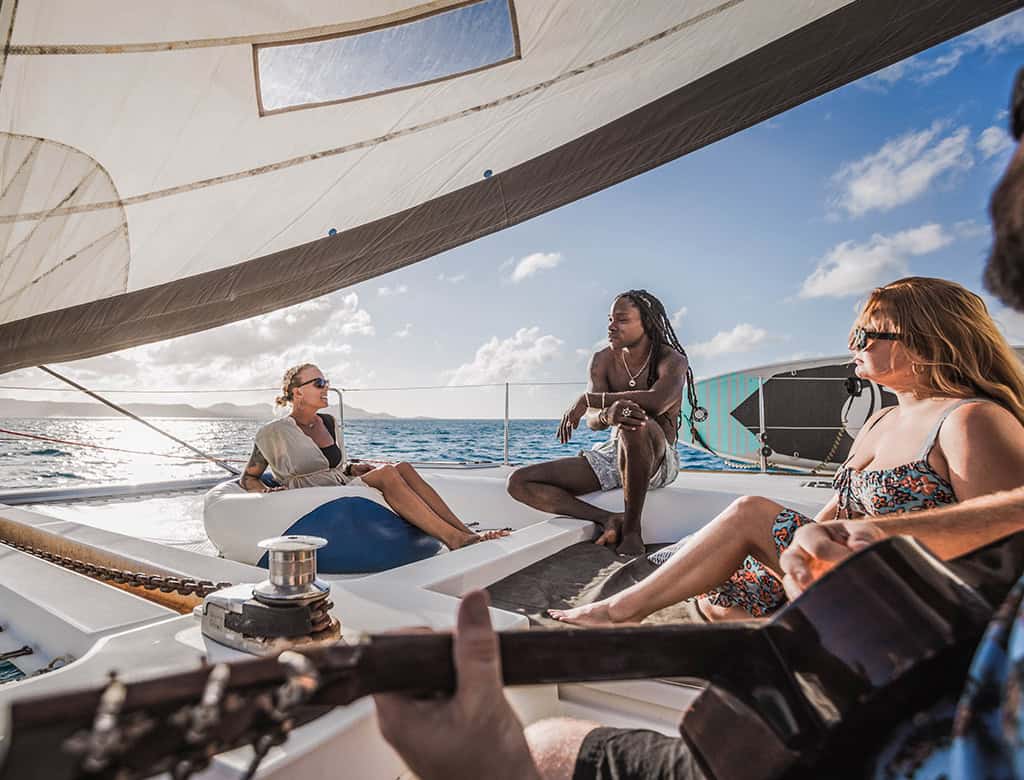 A group of people sit in the sunshine on a sail boat smiling. One person is holding a guitar.