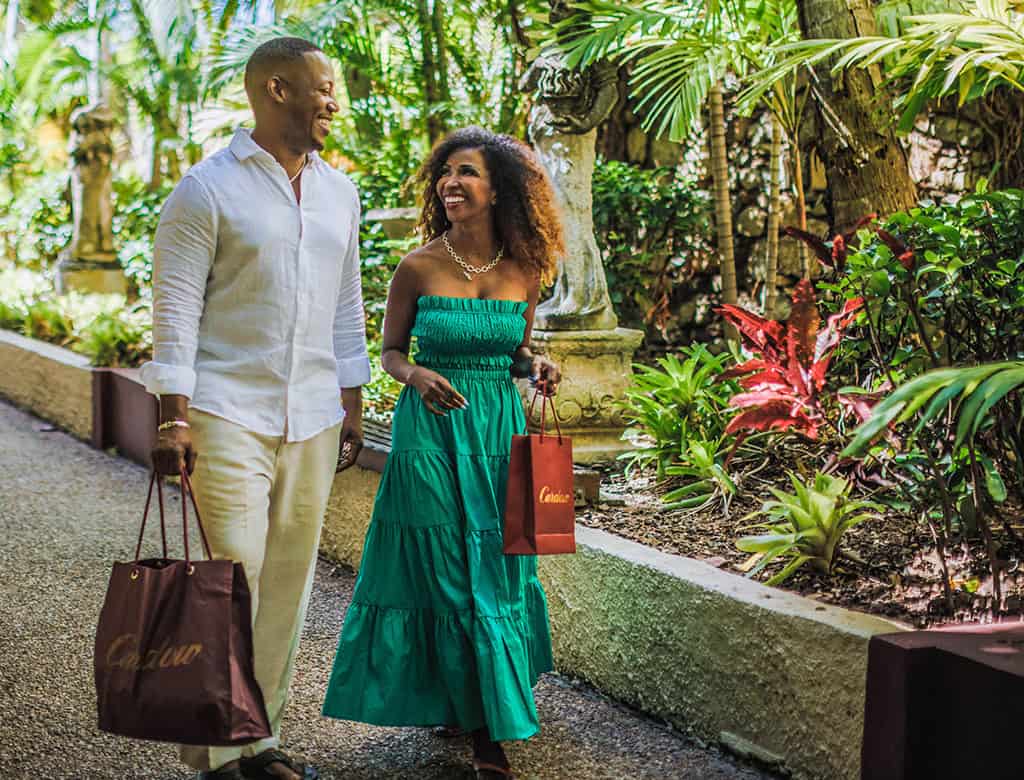 A couple smiles as they stroll through St. Thomas with shopping bags in hands. They are surrounded by tropical plants.