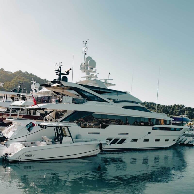 A large white yacht and a smaller boat anchored side by side in a marina with hills in the background.