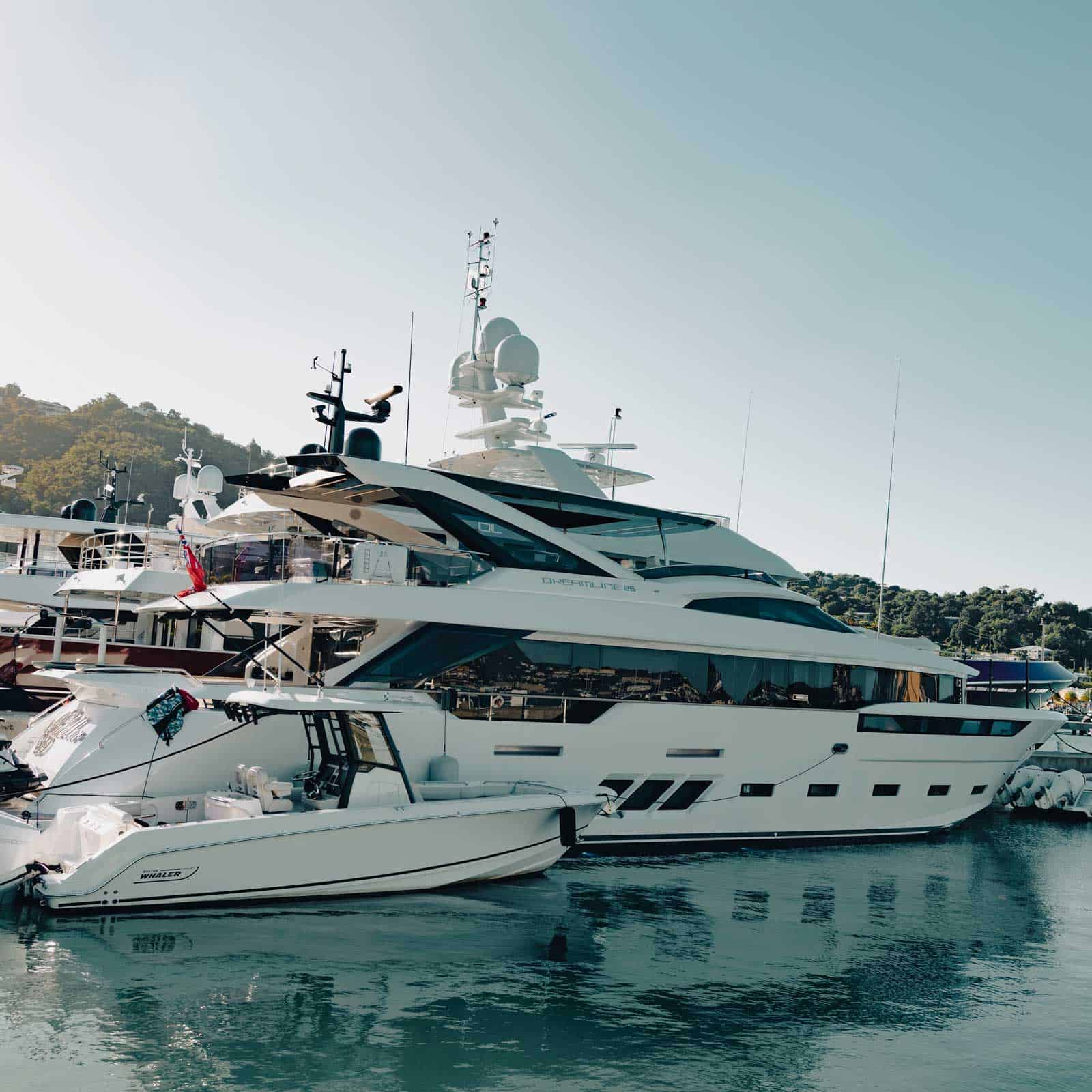 A large white yacht and a smaller boat anchored side by side in a marina with hills in the background.