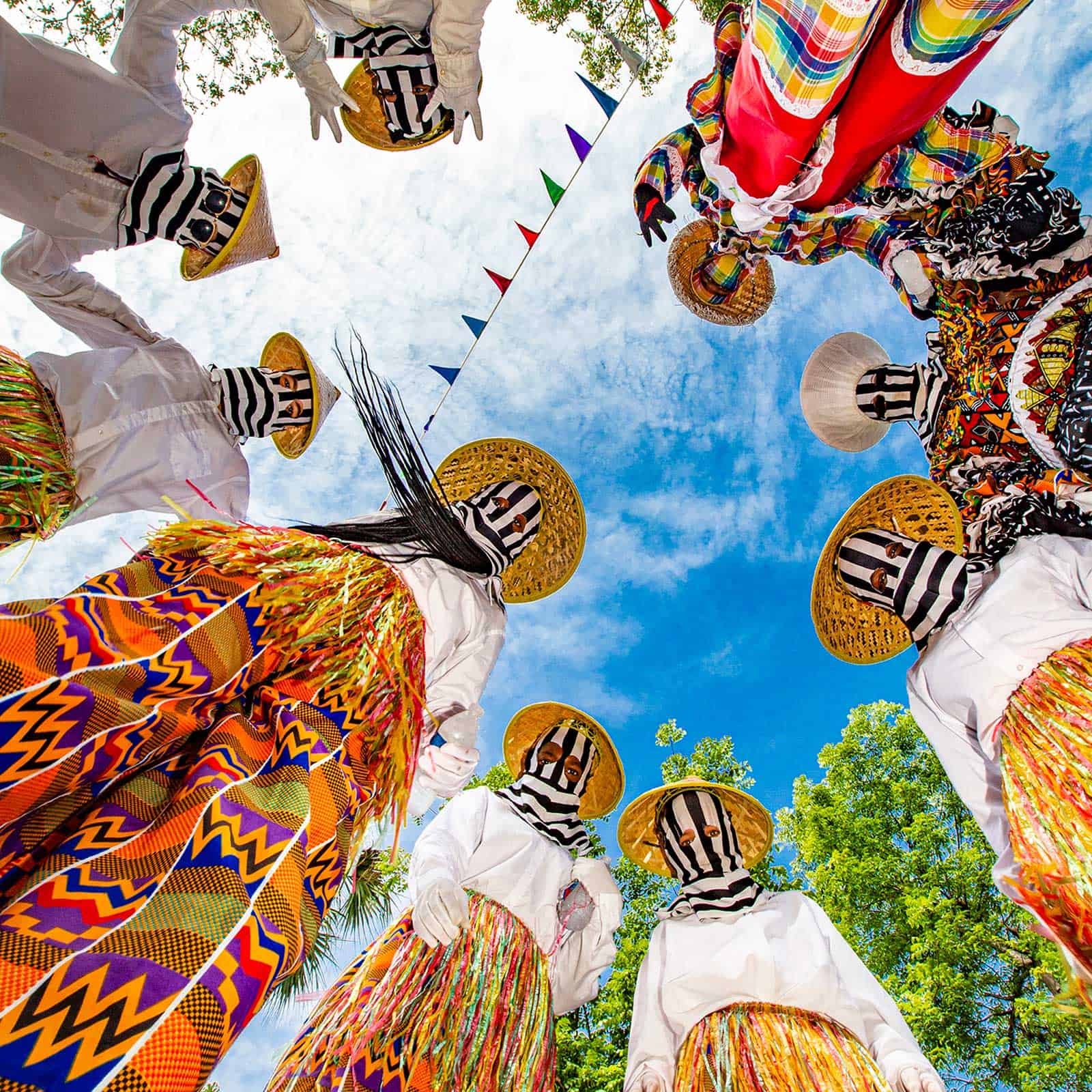 A group of people in colorful costumes and striped masks stand in a circle looking down at the camera, with a bright blue sky and trees in the background.