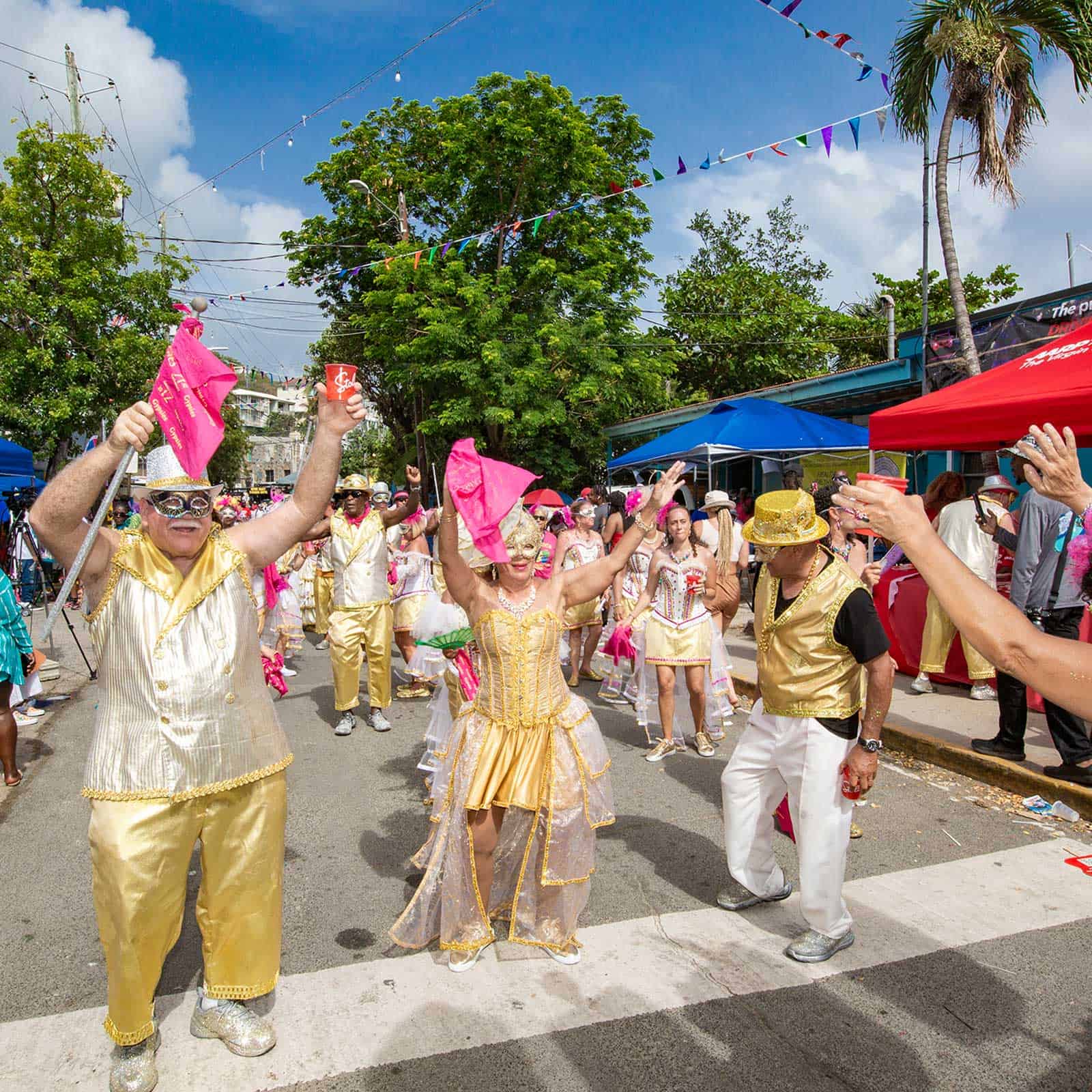 A group of people dressed in gold and white outfits dance and wave pink flags at an outdoor event under a clear blue sky with palm trees in the background.