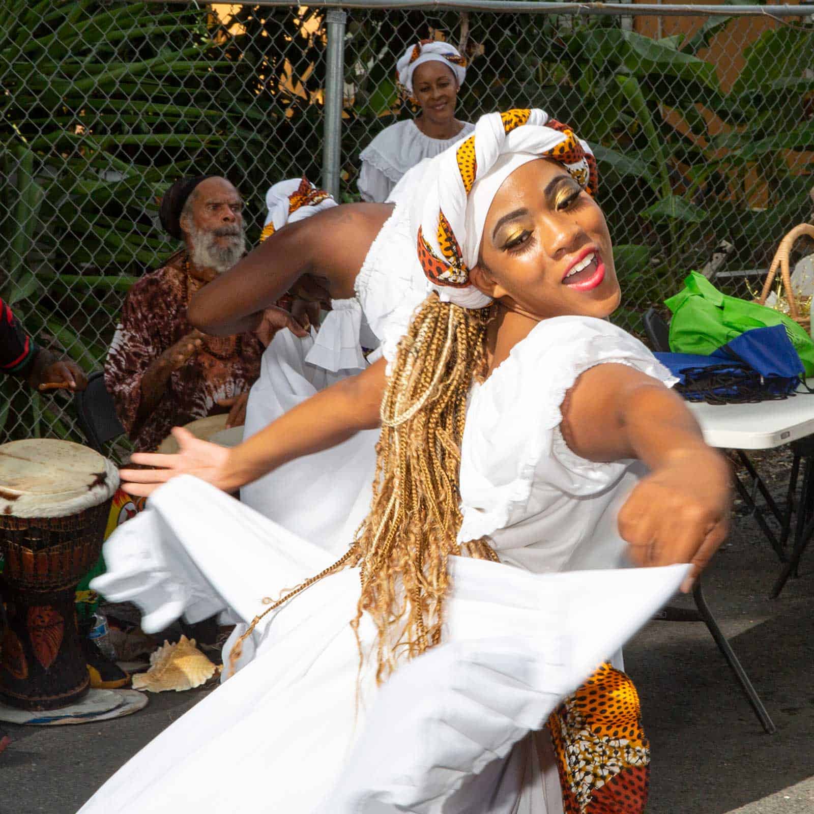 A dancer in a white dress performs energetically outdoors, while musicians play drums in the background. Other people in traditional attire are also present.