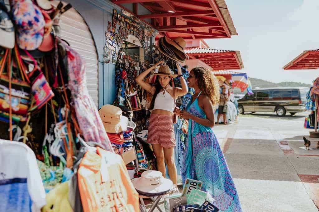 Two women examine hats at a colorful outdoor market stall in Charlotte Amalie. One tries on a hat while the other observes. Various clothing items and accessories are displayed around them.