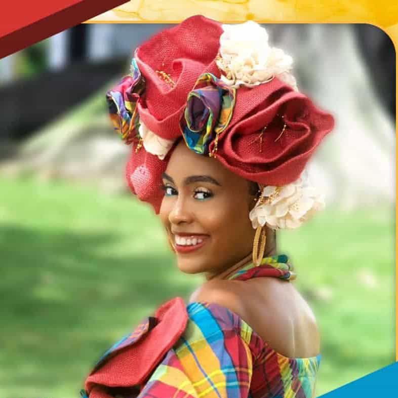 A person smiling, wearing colorful traditional clothing and a large headpiece with fabric flowers, outdoors with blurred greenery in the background.