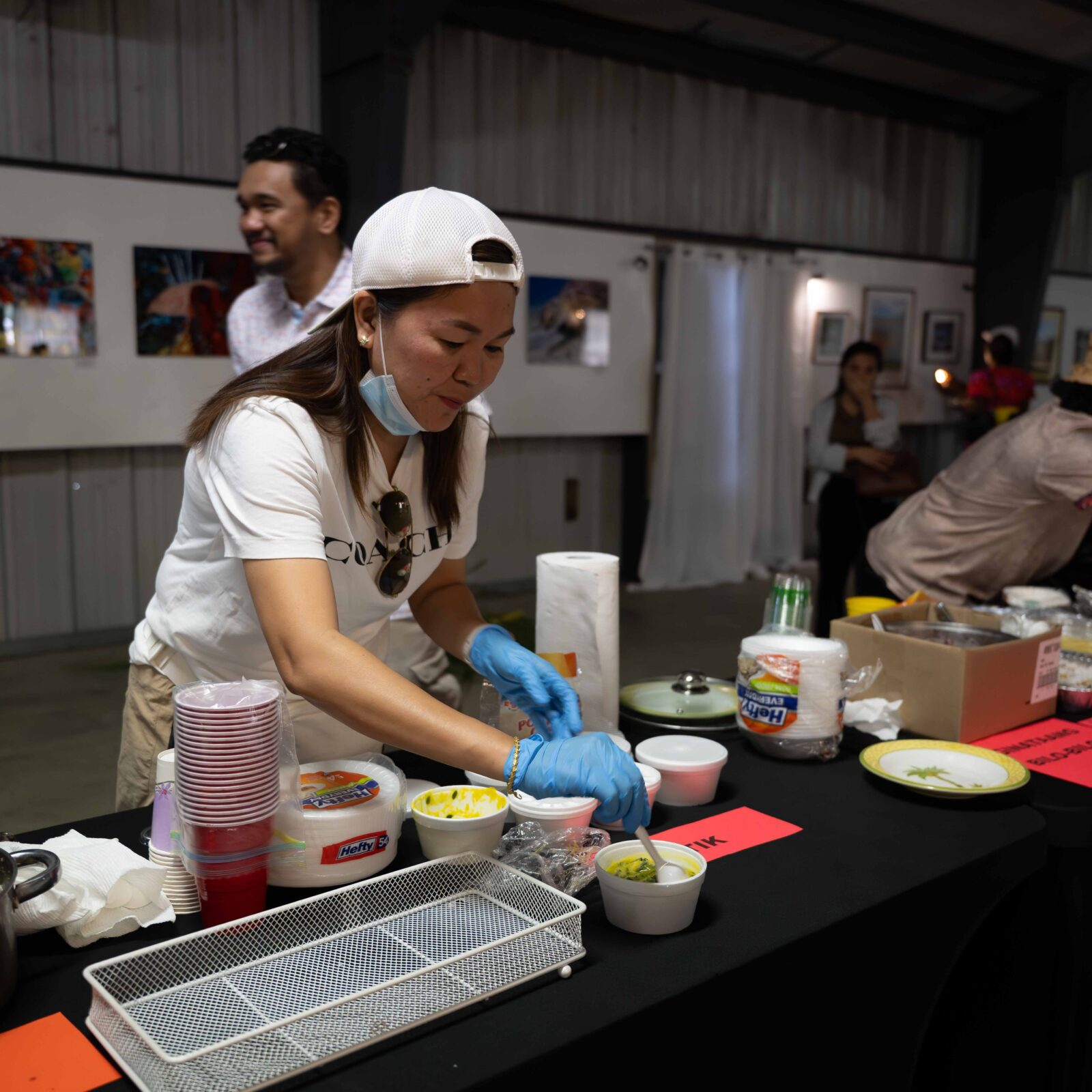 A woman wearing gloves and a cap prepares food at a serving table indoors, with containers, plates, and utensils arranged in front of her at the Crucian Coconut Festival on St. Croix. Other people are in the background. THIS IMAGE CAN BE USED ONLY IN CONJUNCTION WITH THAT FESTIVAL
