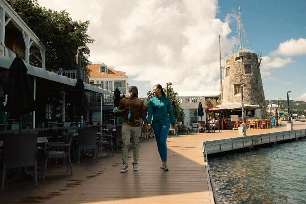 A couple walks hand in hand along a waterfront boardwalk, passing outdoor dining areas and an old stone tower under a partly cloudy sky.