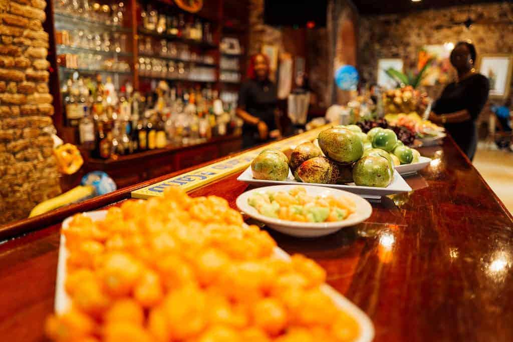 A wooden bar counter displaying assorted fruits and snacks with bottles of alcohol arranged on shelves in the background. Two people are seen in the dimly lit setting.
