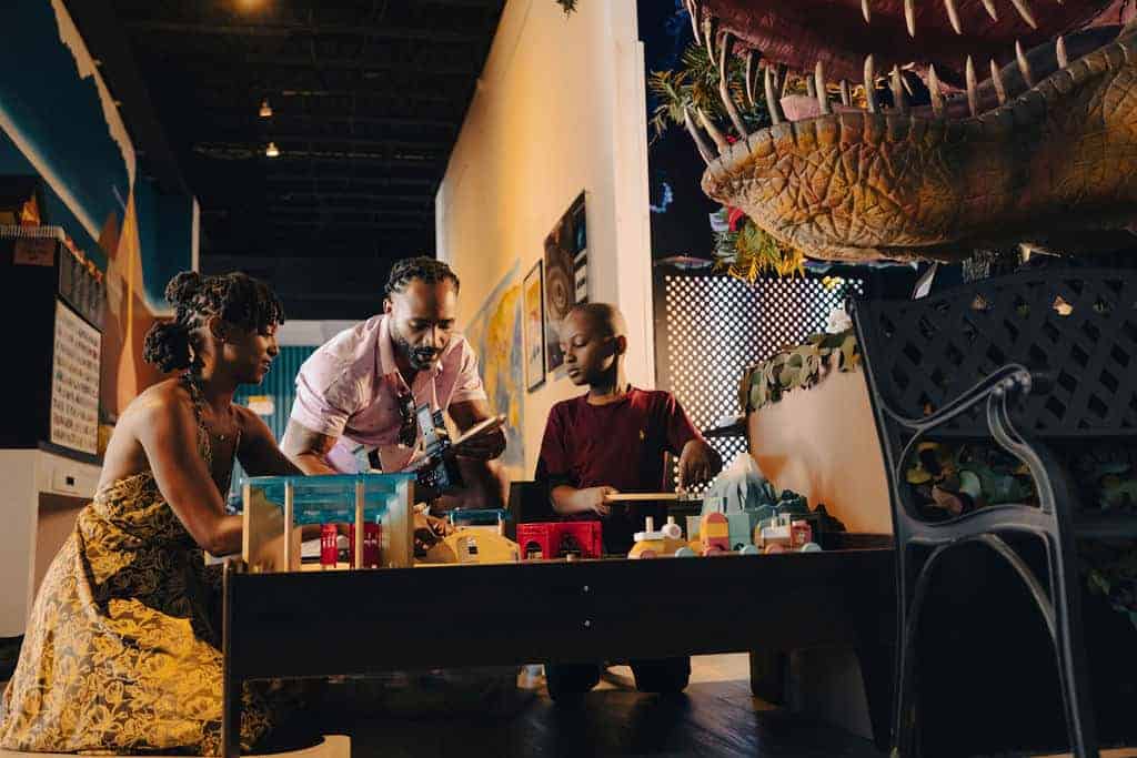A man, woman, and child play with toys on a table under a dinosaur model in a well-lit room.
