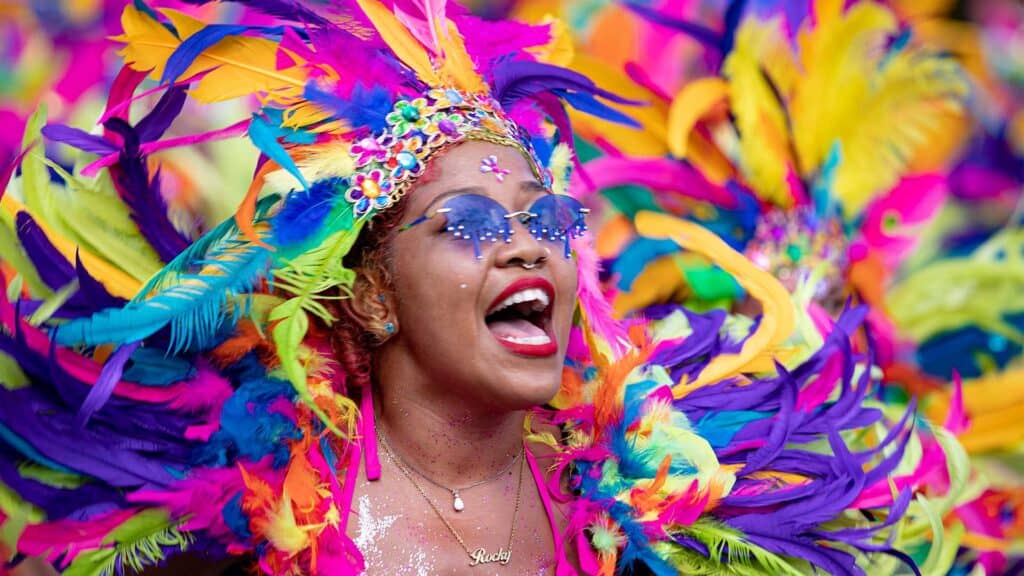 A person wearing vibrant, colorful feathered costume and sunglasses sings joyfully, adorned with flowers and jewelry, against a matching colorful background.
