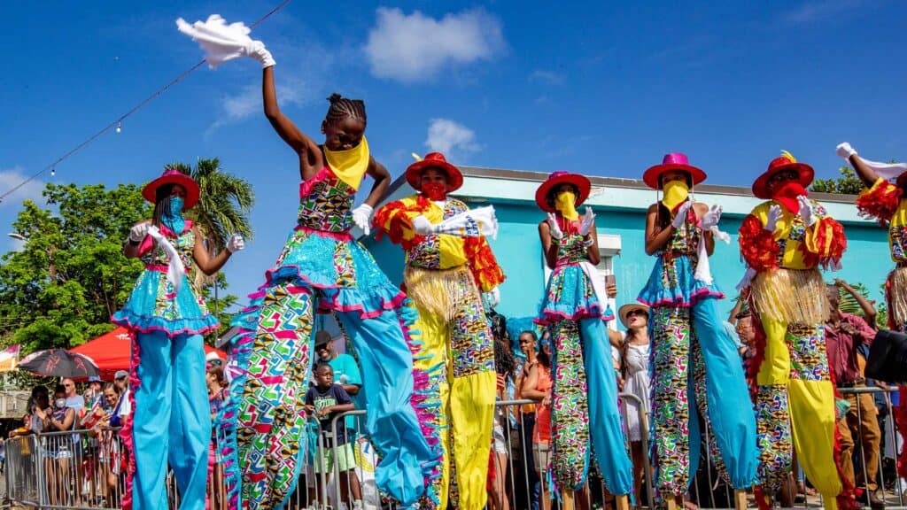 People in colorful moko jumbie costumes and on stilts perform in a lively parade with a bright blue sky and spectators in the background in the USVI.