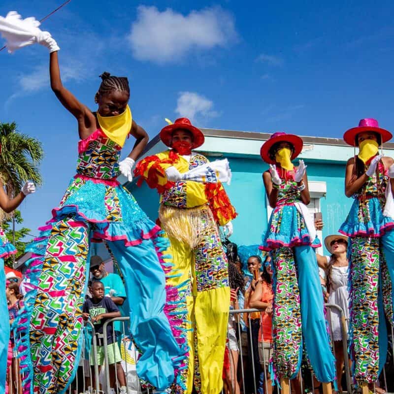 People in colorful moko jumbie costumes and on stilts perform in a lively parade with a bright blue sky and spectators in the background in the USVI.