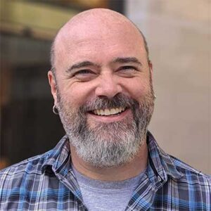A smiling bald man with a beard and wearing a plaid shirt is looking slightly off-camera. He is in an outdoor urban setting.