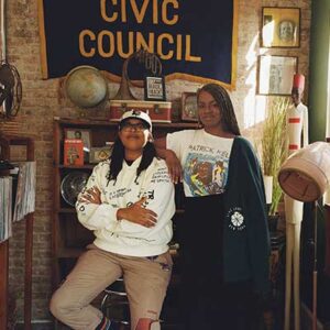Two people posing together in a room decorated with a "Civic Council" banner, globe, books, and various objects. One person is sitting, the other is standing with a hand on the first person's shoulder.