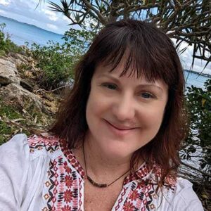 A woman with dark hair, wearing a white blouse with red patterns, smiles at the camera with a backdrop of water, rocks, and greenery.