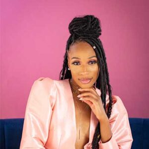 Person with long braided hair and a topknot, wearing a pink satin outfit, posed against a pink background with a thoughtful expression, resting their hand on their chin.
