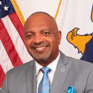 A man in a grey suit and blue tie is smiling in front of an American flag and another flag with an eagle emblem.
