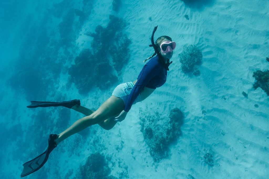 A woman wearing a snorkel and fins swims underwater near the sea floor with scattered rocks and coral.