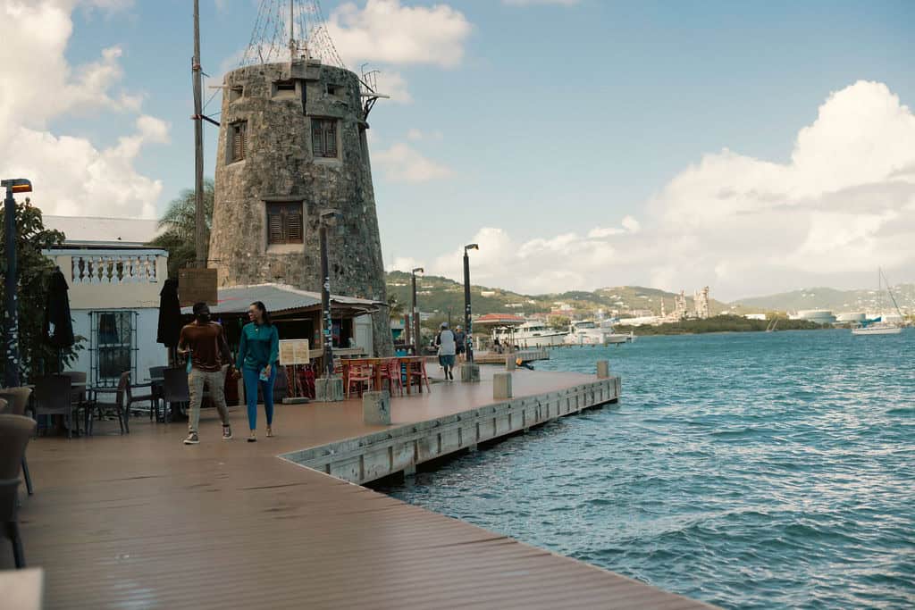 People walk along a wooden boardwalk next to a body of water, with a stone tower and buildings in the background under a cloudy sky.