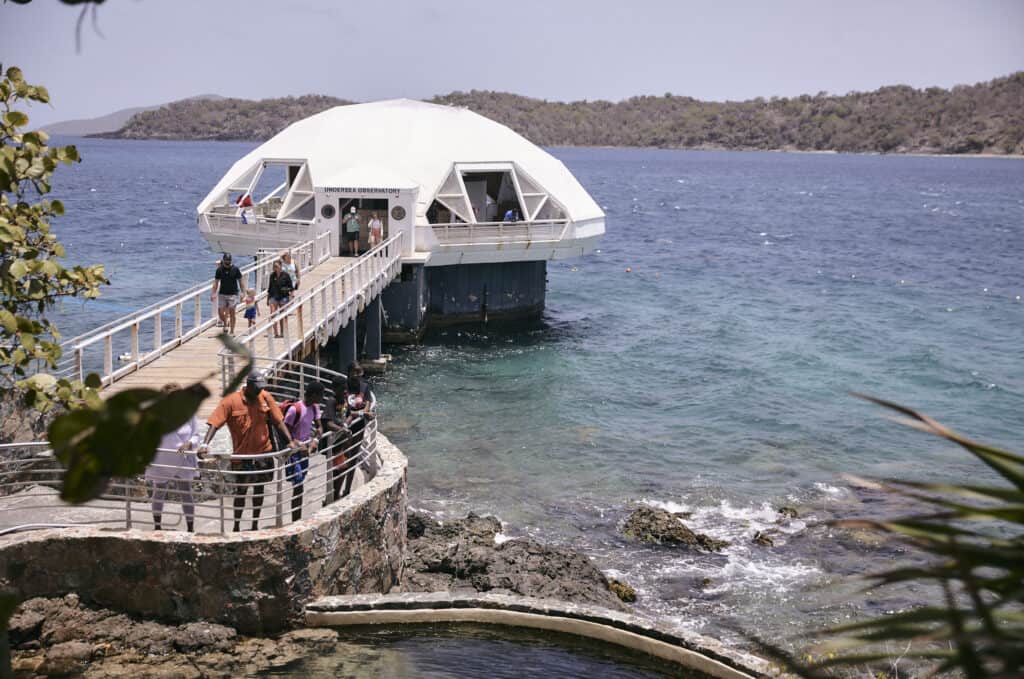 A group of people walks on a boardwalk toward a domed structure over the ocean, near a rocky coastline and distant hills at Undersea Observatory at Coral World Ocean Park in St. Thomas, USVI