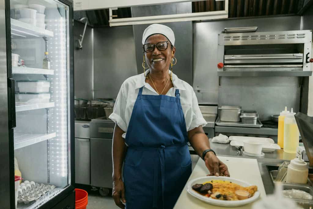 A person in a white shirt and blue apron stands in a kitchen, smiling. They are next to a plate of food on a counter at Miss Lucy's on St. John in USVI