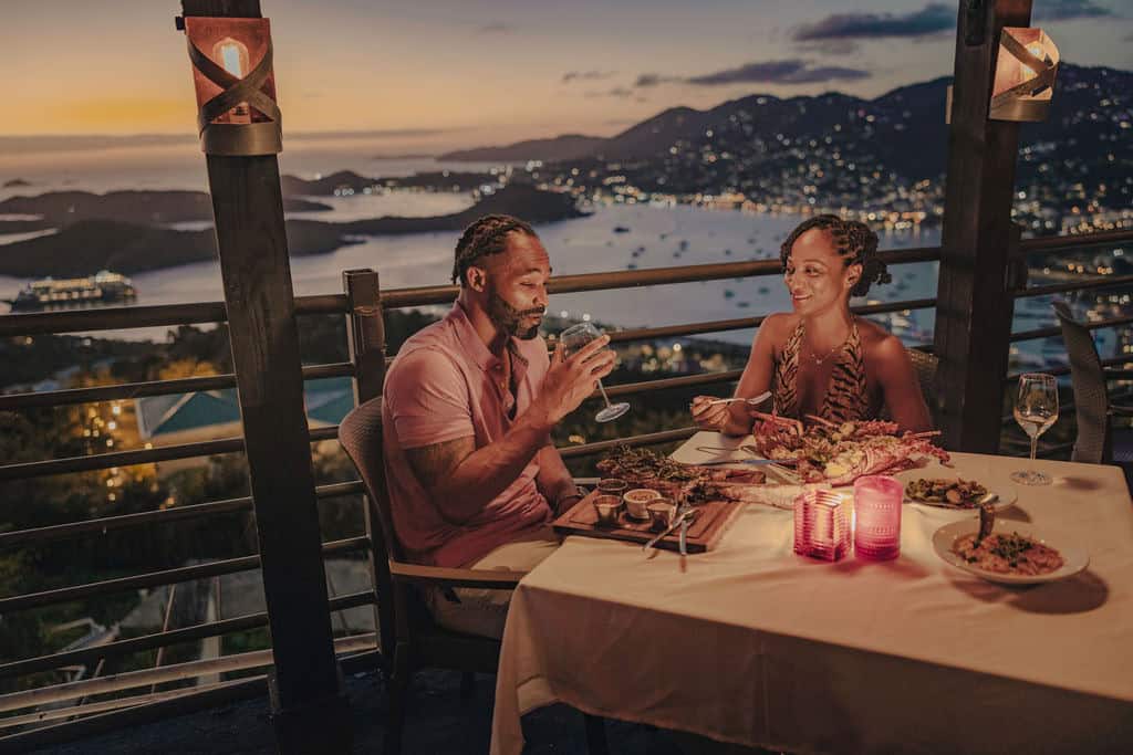 A couple enjoys a candlelit dinner on a balcony with a scenic view of a bay and mountains at sunset at PRIME at Paradise Point, St. Thomas, USVI
