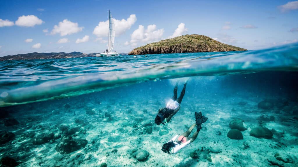Two snorkelers swim near the ocean floor in clear water, with a sailboat and a green island visible in the background above the water's surface.
