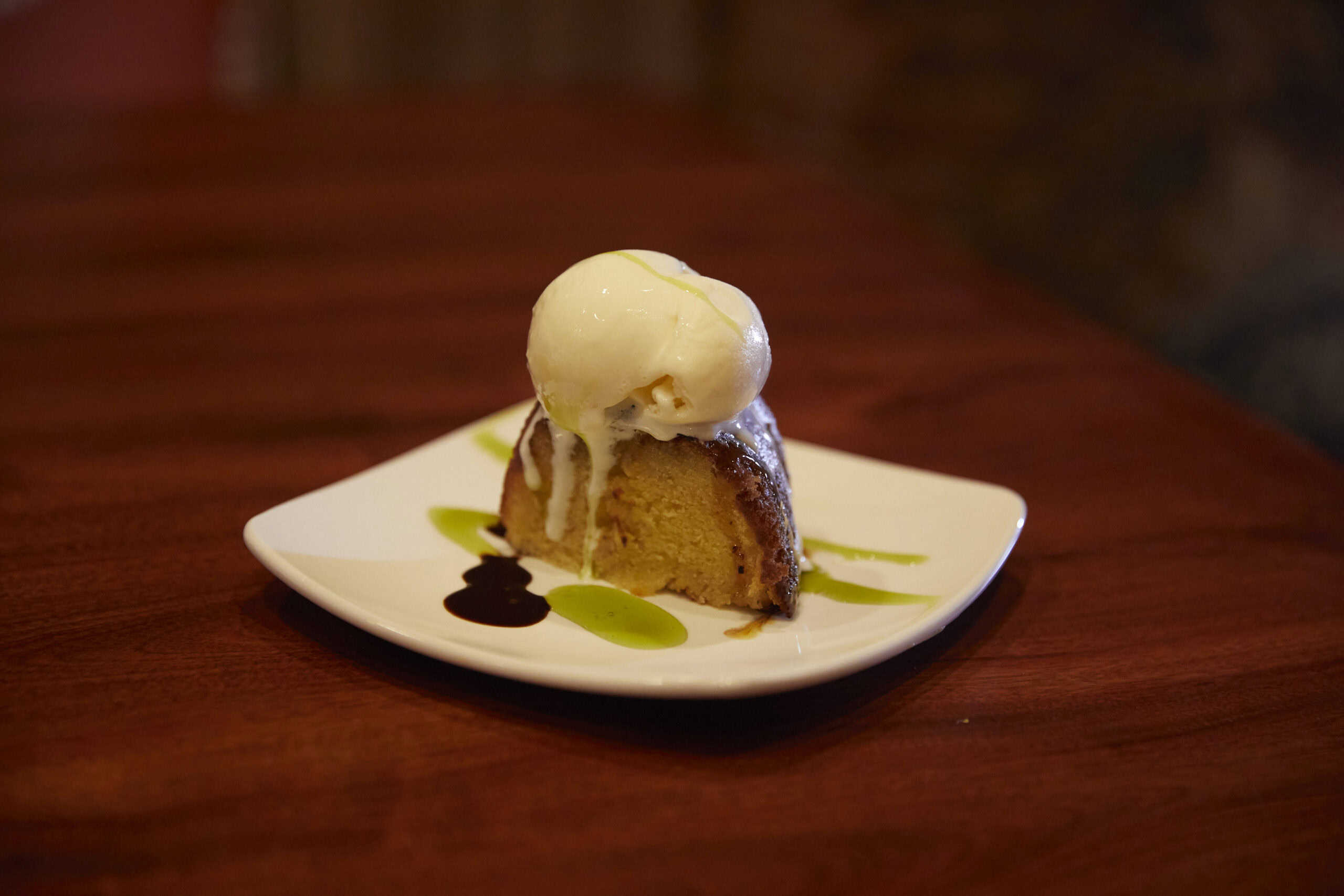 A slice of rum cake topped with whipped cream, served on a square white plate with brown and green sauce drizzles, on a wooden table at Surf N' Turf Bistro in St. Thomas, USVI.