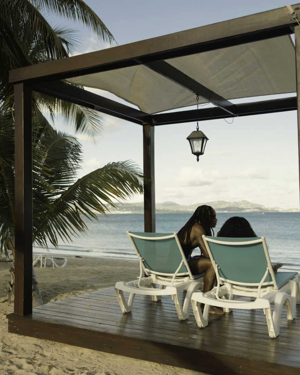 Two people sit on lounge chairs under a beach cabana by the water, with a palm tree nearby and mountains visible across the sea in the background.