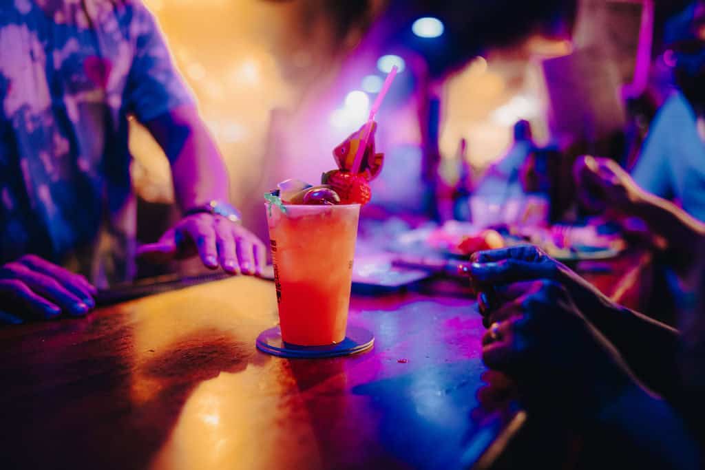 A colorful cocktail with a straw and garnishes sits on a bar counter. Two people are nearby in a dimly lit setting with vibrant purple and orange lighting.