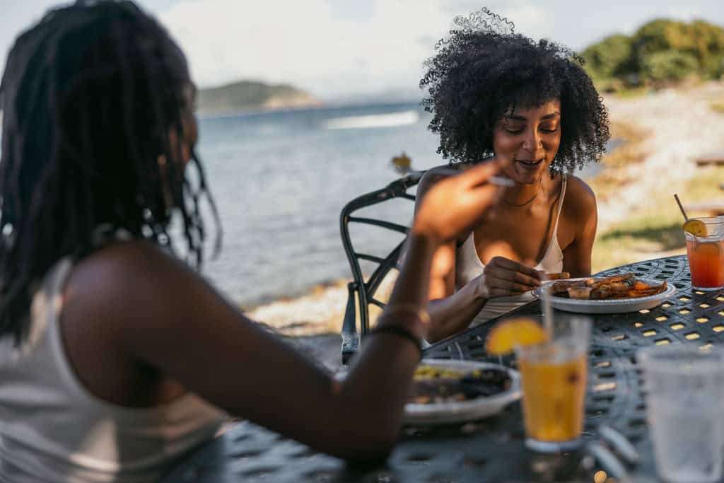 Two people sit at a metal outdoor table by the water, enjoying meals and drinks in a sunny setting at Miss Lucy's on St. John, USVI.