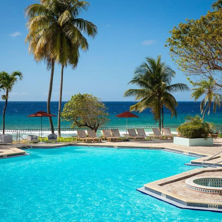 Swimming pool with lounge chairs, palm trees, and umbrellas by the ocean. Clear blue sky in the background.