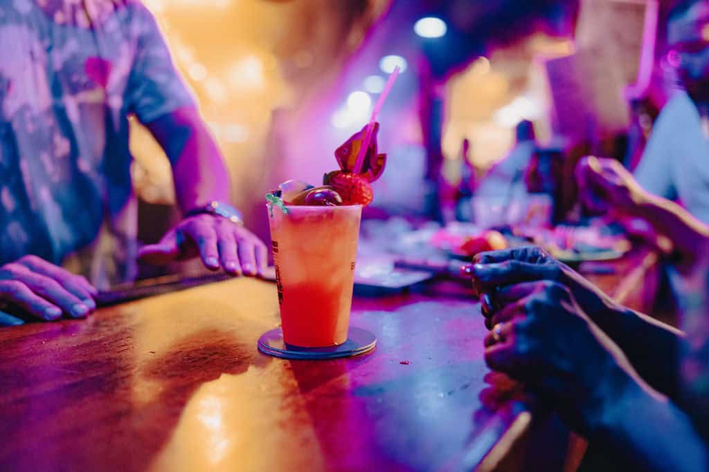 A vibrant cocktail with a straw and garnish sits on a bar counter, surrounded by people in a lively, colorful setting.