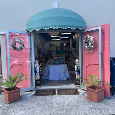 Storefront with open pink double doors, green awning, and decorative wreaths. Two potted plants and wooden figures stand at the entrance. Inside, a round table with items is visible.