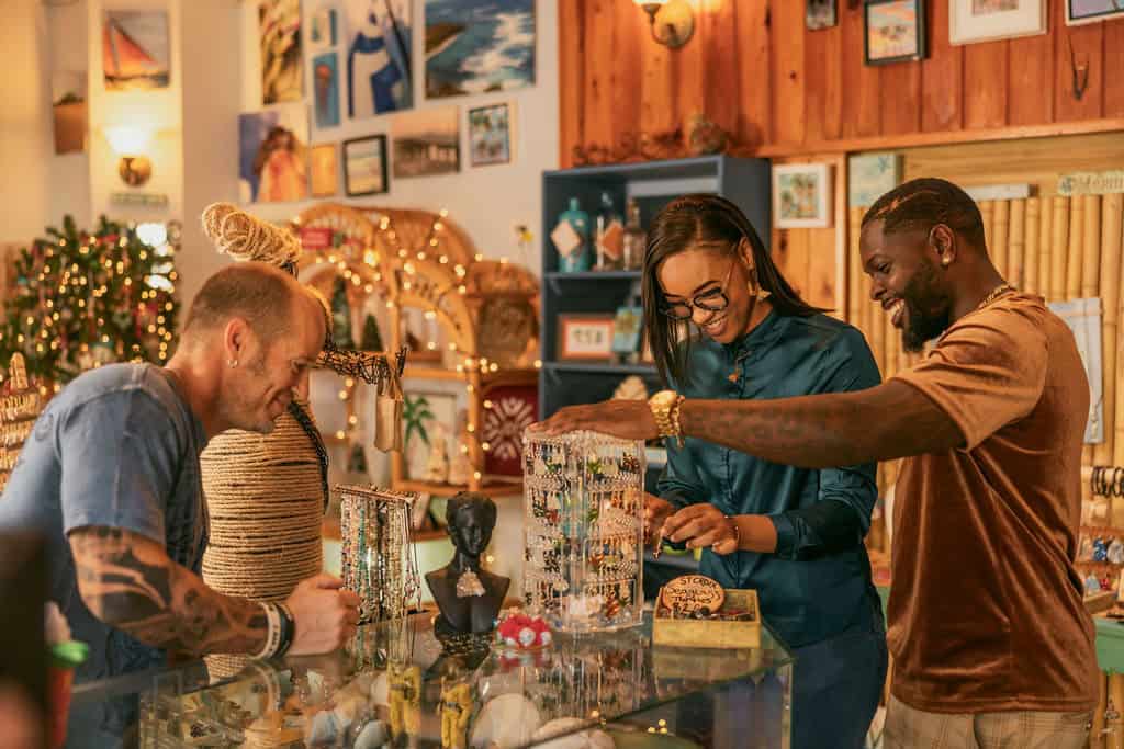 Three people browse jewelry in a warmly lit store with wooden walls in Christiansted, St. Croix, U.S. Virgin Islands. Various items and artworks are displayed around them.