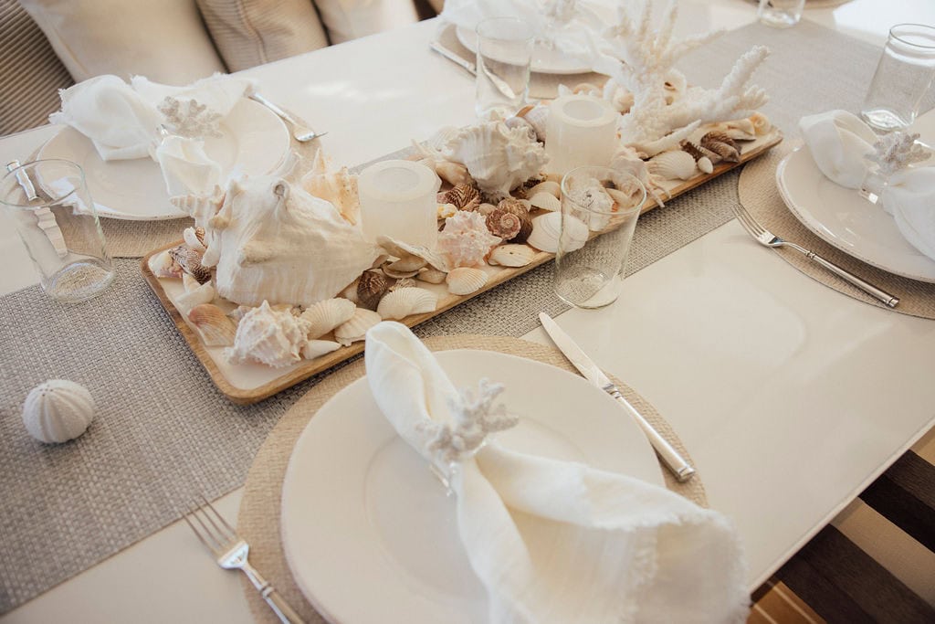 Table setting with white plates, napkins, and glasses. Centerpiece features seashells and corals. Neutral-toned placemats and table runner complete the coastal-themed decor in St. Thomas, U.S. Virgin Islands.