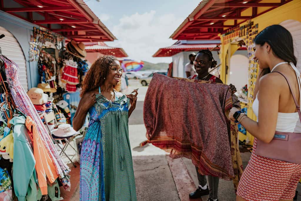 Three women in the Vendors Plaza marketplace along the Charlotte Amalie waterfront in St. Thomas, U.S. Virgin Islands. Two hold up a colorful dress. Bright stalls display clothing and accessories. Sunny daytime setting.